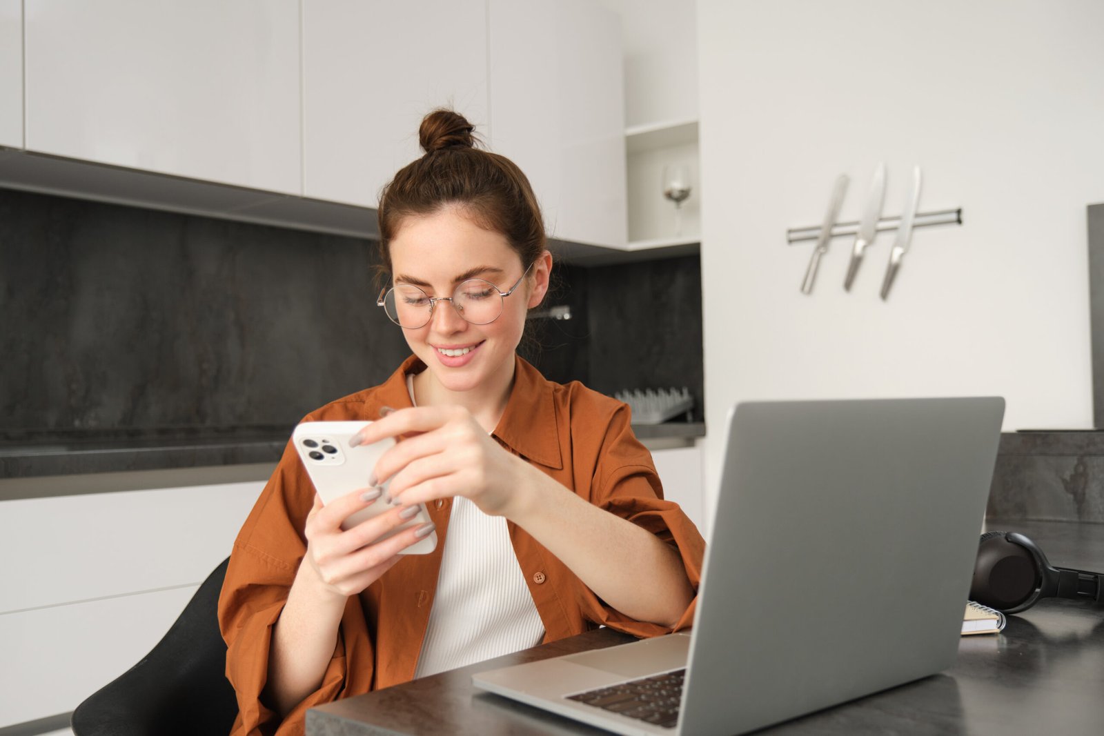 Portrait of young female freelancer, woman working from home with laptop, girl programmer sitting with computer in kitchen, answer message on smartphone.